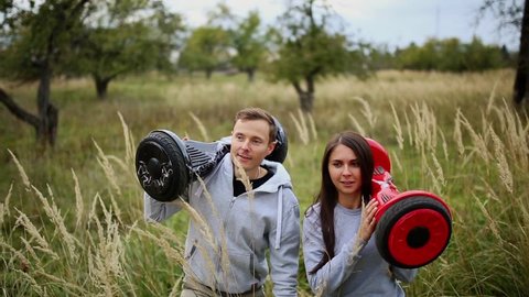 Young man and girl go into the field with hoverboards look at one another in urban clothing. Steadicam