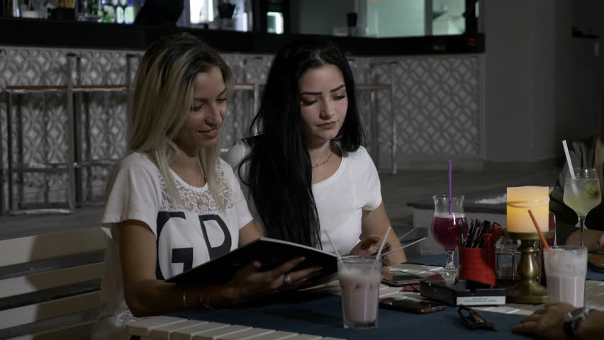 Two young beautiful girls in club sitting at a restaurant table trying to decide what to order from the menu