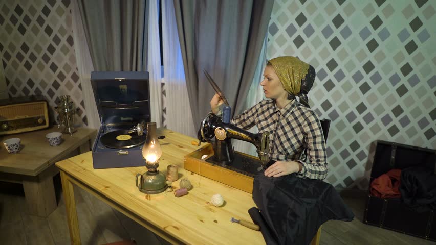 Beautiful retro seamstress girl sews cloth with old manual hand sewing machine. Woman working at home or workshop at night with kerosene lamp, listens music on vinyl plate, gramophone or phonograph