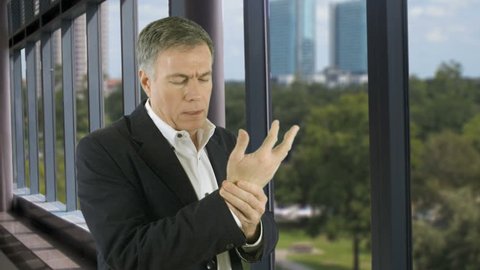 A concerned man in a high rise office building stands by the windows and holds his wrist as if in pain.