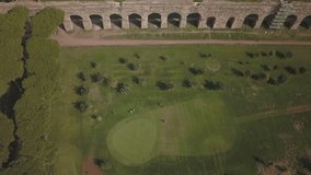 Video from above. Aerial view of people playing golf in a field near the Roman Aqueduct.	