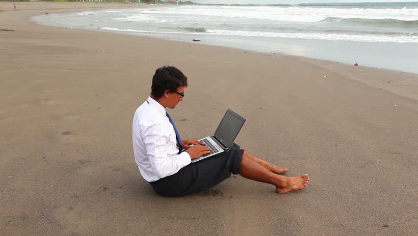 young guy in office suit printing something on laptop sitting on the beach