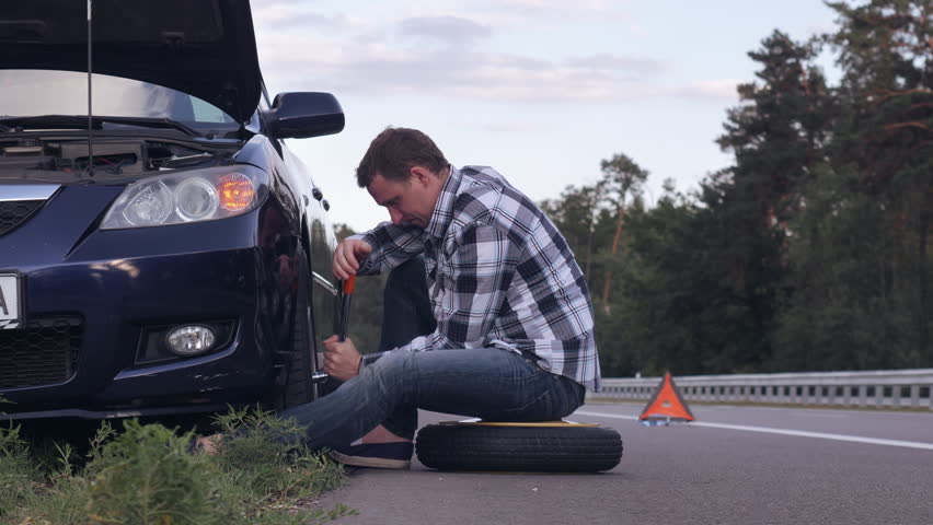 Caucasian man changing a tire sitting on stepney near car on the roadside. Handsome traveller outdoors