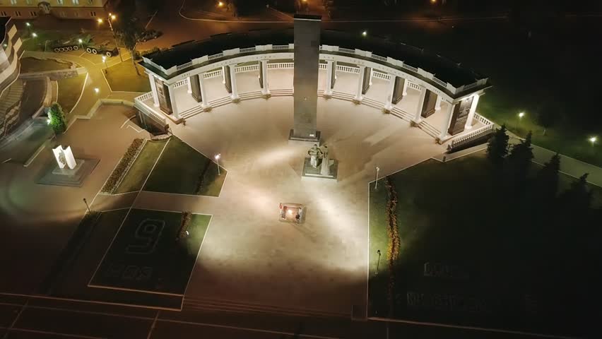 Memorial to the soldiers of Mordovia who fell during the Great Patriotic War. The memorial complex on Victory Square in Saransk features a tall central granite obelisk set before a sweeping semicircular colonnade with evenly spaced columns and balustrades. A bronze sculptural group of a woman and soldier stands at the base of the obelisk, while an eternal flame burns in a low star-shaped platform at the center of the paved plaza. The complex is seen from a high aerial perspective at night, with warm ground lighting illuminating the white stone architecture against the dark surroundings. The adjacent Museum of Military and Labor Feat, with its distinctive facade of multicolored granite slabs in the colors of the St. George