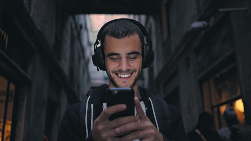 Smiling and laughing handsome young man listening music from his smartphone in wireless black headphones, dancing on street of old town center in europe at twilight or evening time Royalty-Free Stock Footage #32030479