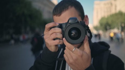 Young male tourist or photography student photographs into camera with big DSLR, clicks shutter to create memories. he smiles and laughs during summer vacation trip in europe