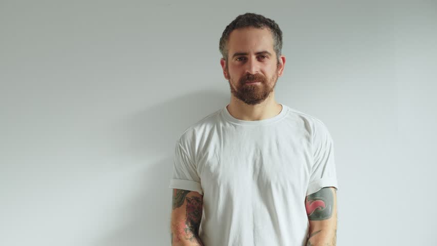 Attractive and handsome middle aged hipster man with cool arm tattoos looks into camera, stands on isolated white wall, girlfriend, spouse or partner pretty woman appears from back, hugs him on neck