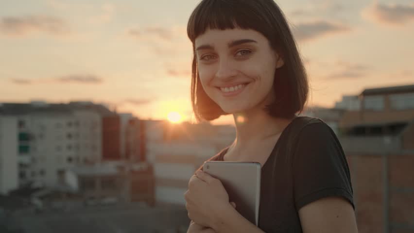 Playful beautiful young woman in her 20s, with short haircut and trendy hipster tattoos stands in warm summer sunset light, holds tablet device, chuckles and smiles into camera, shows tongue