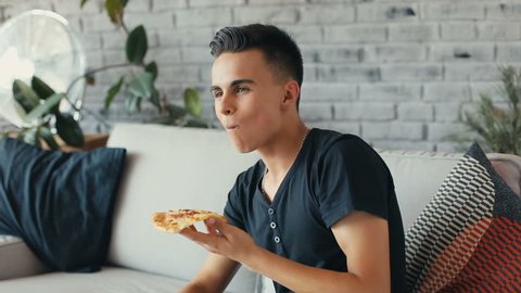 young guy sitting on the couch at home eating pizza smiling. Shot on RED Epic Camera. Slow motion