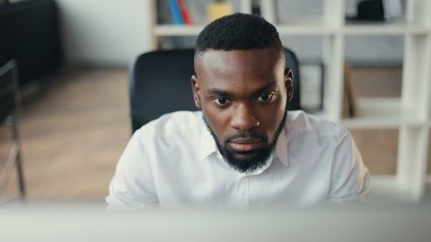 African American handsome male uses computer for work in modern office programmer serious looks at monitor screen freelance successful worker businessman solves engineering problems Slow motion