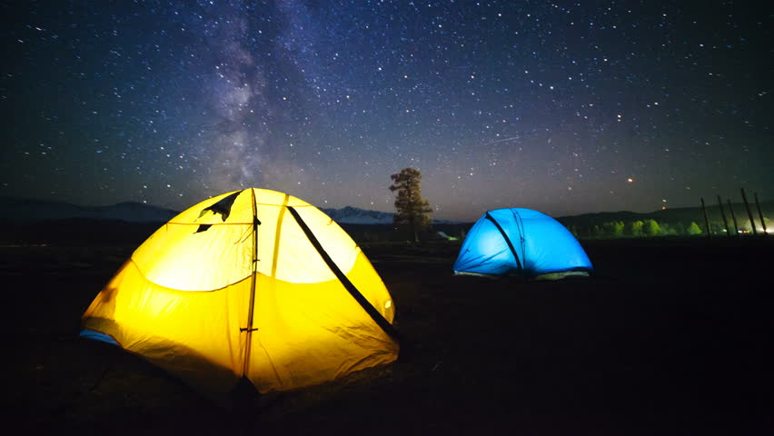 Two travelers watching the meteor shower in summer time. Timelapse of stars moving in night sky over the camp tent