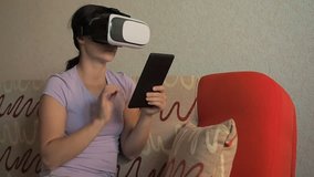Beautiful young girl sits on couch with virtual reality glasses. New technologies. Young excited woman wearing VR headset touching objects in virtual world, using goggles mobile application smartphone