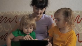 Pretty little girl, boy and mother sitting on sofa using laptop. An adorable family sit together and play with a laptop and digital tablet. 