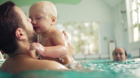 Cute baby having swimming lesson with his mother and father. Healthy family teaching their baby to swim in the swimming pool. Young father takes his son and embraces him while the kid is smiling
