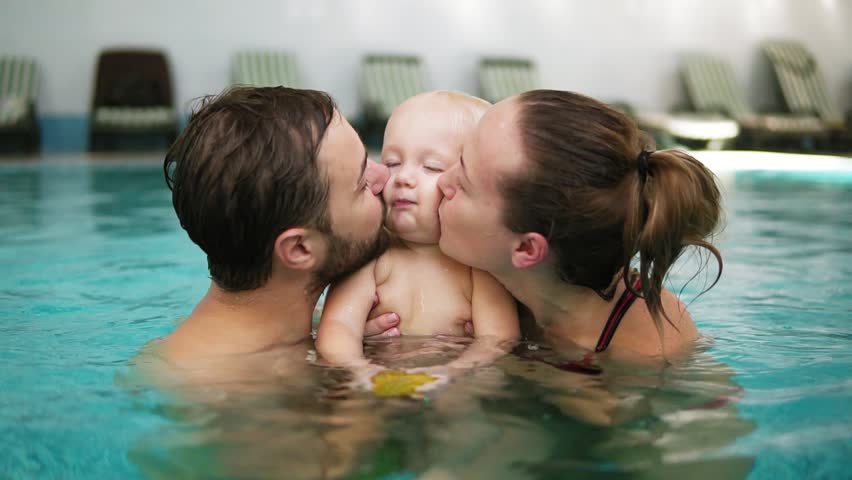 Young mother and father are holding their cute child in their hands in the swimming pool and kissing him from both sides. Happy child is smiling