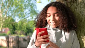 4K video clip of beautiful happy mixed race African American girl teenager leaning against a tree with a red backpack and using a cell phone camera to take pictures and social media