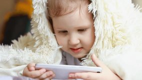 Child Looks cartoons and plays downloaded application on a smart phone close-up. A little cute funny girl lies in bed under the blanket, smilling and looks at the white phone screen.