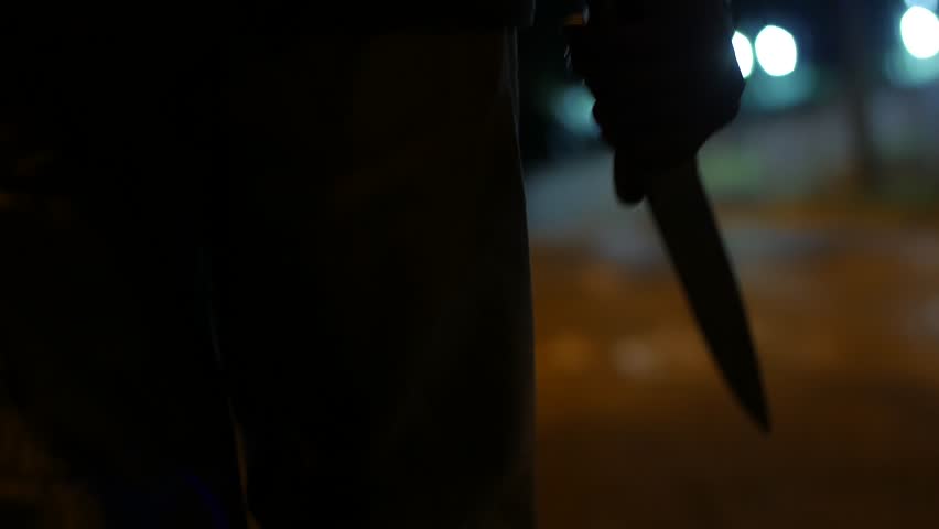 A MAN WALKS AT NIGHT HOLDING A BIG KNIFE, BLURRED BACKGROUND, WAIST AND HAND SEEN