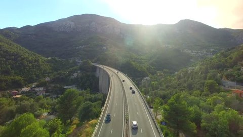 Cars move at highway bridge road in mountains above valley village morning aerial 4k. Fly above traffic on bypass over hill town Europe sun shine. Motorway vehicles pass scenic Italian Alps landscape