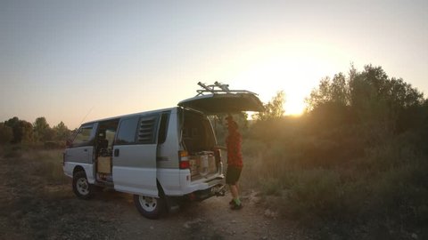 Camera follows handsome trendy hipster man on adventure road trip, he closes trunk of camping van, shuts side door and sits into driver seat, ready to drive through gravel off road
