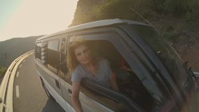 Beautiful young woman with long blonde hair hangs out of camping van window of car, holds selfie stick or camera stabilizer and makes video of adventures. slow motion happinness outdoors