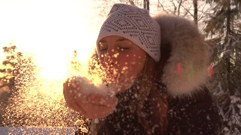 SLOW MOTION CLOSE UP PORTRAIT: Happy young woman blowing white snowflakes into camera at golden winter sunset. Pretty girl sending snowy kiss on cold winter day. Woman in winter clothing blowing snow