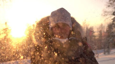 SLOW MOTION CLOSE UP: Young woman blowing snowflakes over the setting sun in winter. Smiling girl sending a snow kiss on gorgeous sunny day on winter evening. Playful girl blowing snowflakes at sunset