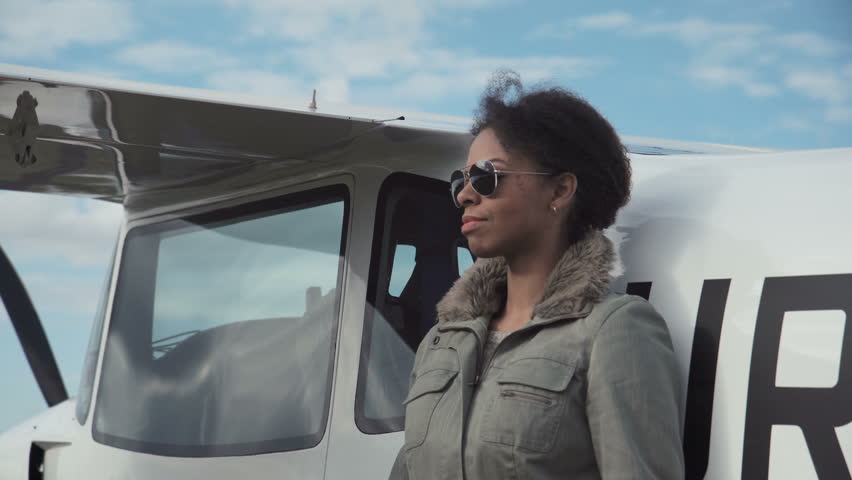 Attractive woman pilot wearing sunglasses standing in the sunshine on an airfield resting against her small private airplane