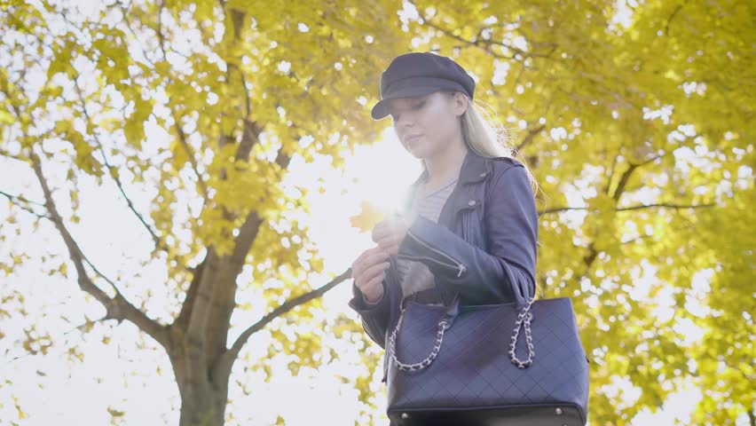 thoughtful pretty blonde is strolling in a park in autumn day, taking yellowed maple leaf near chest, tilt up shot