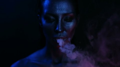 Halloween Vape Party – Nightlife. Beautiful Sexy Young Woman with glamorous mystical makeup vaping in Nightclub (exhaling smoke). Girl smoking vaporizer in Club. Blue mystic smoke