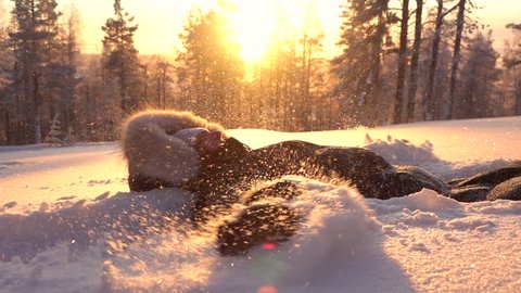 SLOW MOTION Happy girl in warm winter clothing doing snow angels in fresh powder blanket. Cheerful woman laying in powder snow, making angle at sunrise. Playful girl enjoying winter, snowflakes flying