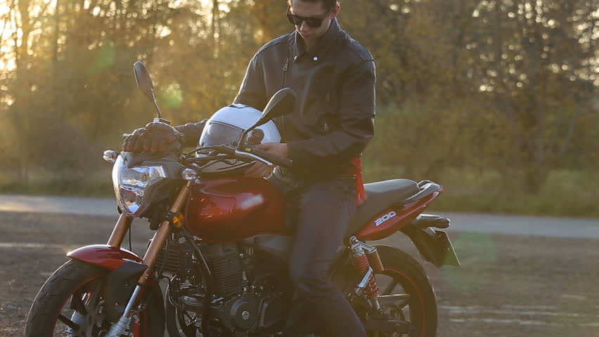 A young man in black leather jacket with white helmet goes to his motorcycle and sits on it before journey at autumn sunset. Close up shot. | Shutterstock HD Video #32129239