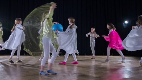 A girl with a big blue ball is dancing in the middle o a stage and the rest of the girls are dancing around her.