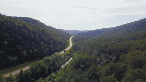 AERIAL: Trucker moves among wild mountain forest. Camera fly above green trees to the curvy road behind semi-truck. Cargo transportation, nature landscape in sunny moment of summer.