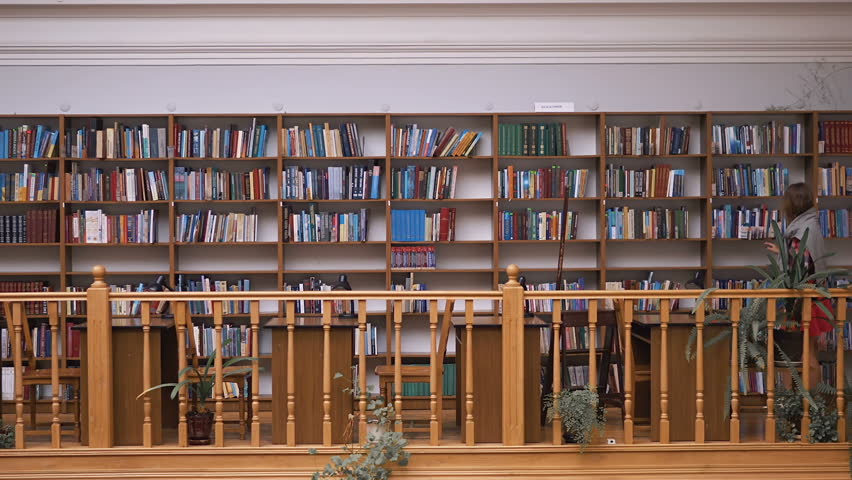 Young casual student walks in library along bookshelves wall