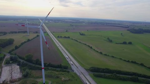 Fly close to wind turbine propellers spinning above road aerial 4k. Wind farm power station in Europe produce renewable sustainable energy in distance from car truck lorries move countryside highway