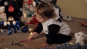 cheerful little girl draws a drawing for Santa Claus near a beautifully dressed Christmas tree. Beautiful Christmas tree with lights. Candles near the Christmas tree