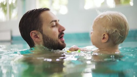 Young father is lifting his little boy from the water while teaching him how to swim in the swimming pool. Happy little boy and his father are laughing and having fun