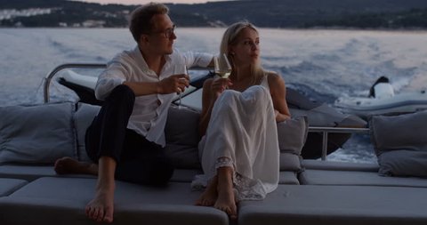 Young Couple Talk, Drink Champagne in the Stern of the Moving Yacht. They Have Great Romantic Evening. In the Background Island with Small Village. Shot on RED Epic 4K UHD Camera.