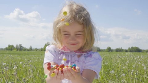 Cute little girl with pleasure catches multicolored candy falling from above. Joyful cheerful child laughing outdoors. Summer sunny day. Slow Motion.