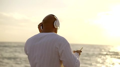 big, bald man in glasses listening to music on my Bluetooth wireless headphones from mobile phone and dances on the background of the sea and sunset. successful middle-aged man on holiday