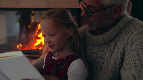 Little girl reading book with grandfather near fireplace at home. Senior man with grandaughter reading a book.
