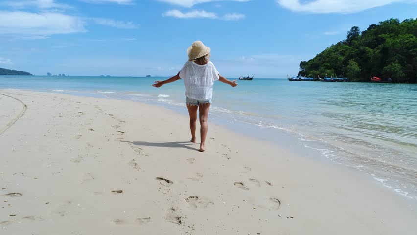 Young woman walking on the beach in Thailand, Drone shot of female walking towards the ocean on a beautiful sunny day, arms outstretched 