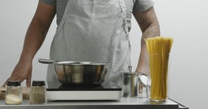 Muscular dark skinned chef in a gray apron adds salt to boiling water and mixes with a wooden spatula on white background