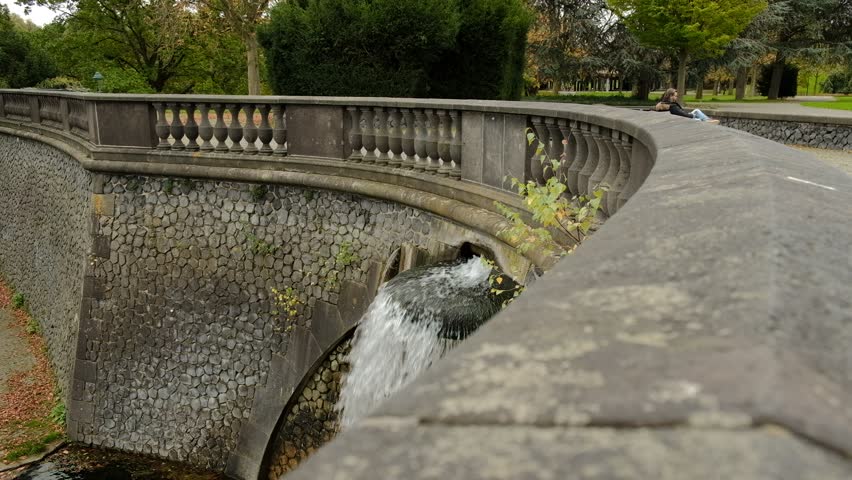 Urban oasis: serene stone structure arches over placid stream in lush park setting under overcast sky. Cascading waterfall creates gentle auditory backdrop with soothing splash against stones. Vibrant foliage displays diverse shades of green surrounding architectural elements. Fallen autumnal leaves introduce warm tones to cool palette, enhancing seasonal atmosphere. Relaxed individual reclines on stone railing, embodying contemplative mood amid harmonious blend of constructed and organic elements. Natural illumination accentuates textural details of materials and vegetation, cultivating peaceful solitude and connection with outside environment. Composition showcases elegant balance between urban infrastructure and natural charm, ideal for lifestyle content, metropolitan exploration, and wellness concepts reflecting moments of stillness and aesthetic appreciation.