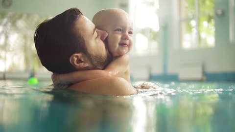 Cute little baby and his father having swimming lesson in the pool. The father is holding his son in his hands and embracing him. Little boy is happily smiling Arkivvideo