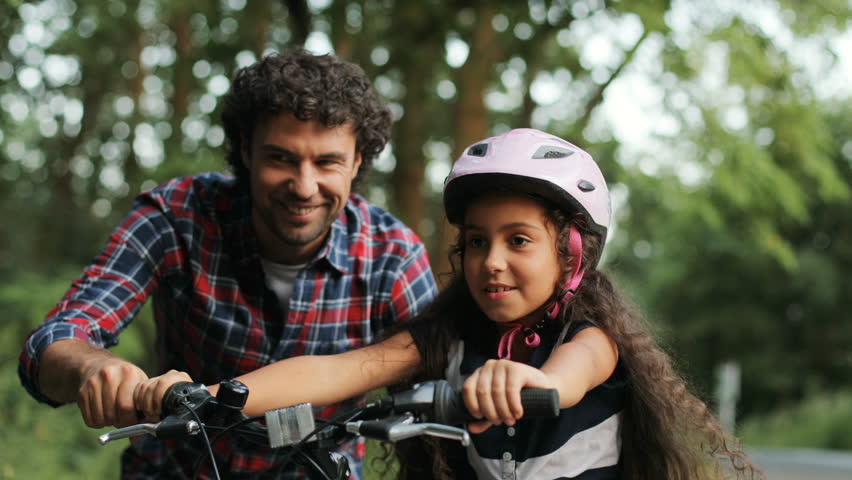 Closeup. Portrait of a little girl. Her dad is teaching her to ride a bike. He lets her go. Happy girl. Moving camera. Blurred background | Shutterstock HD Video #32235646