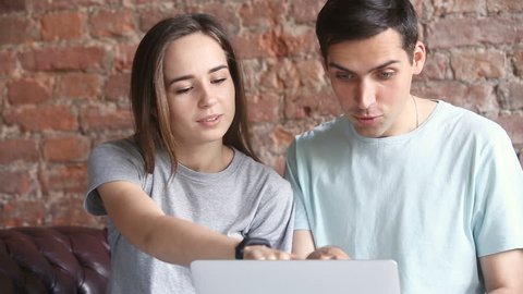 Young casual couple using laptop sitting on couch at home, choosing best sale offers, purchasing goods in online shop together, searching cheap travel tours, booking hotel abroad, comparing prices