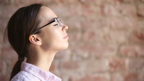Young woman wearing glasses inhaling and exhaling fresh air, taking deep breath, enjoying practicing breathing pranayama exercises indoors, calming down, reducing stress, close up face side view