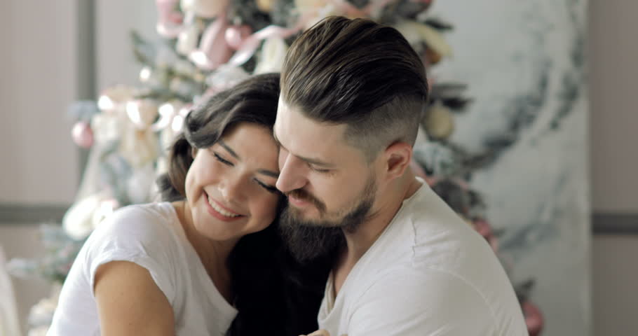 young beautiful couple sitting on a bed next to a Christmas tree, hugging, kissing and laughing